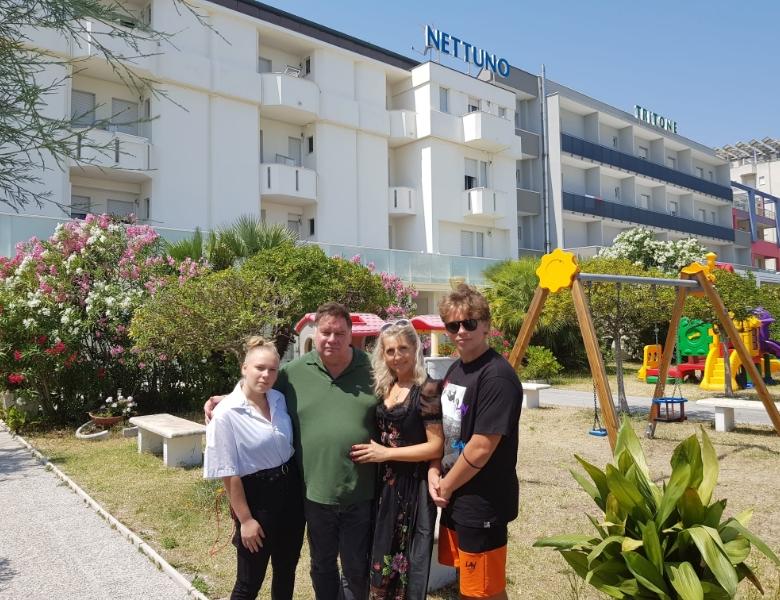 Famiglia sorridente in giardino fiorito, davanti a hotel bianco soleggiato.