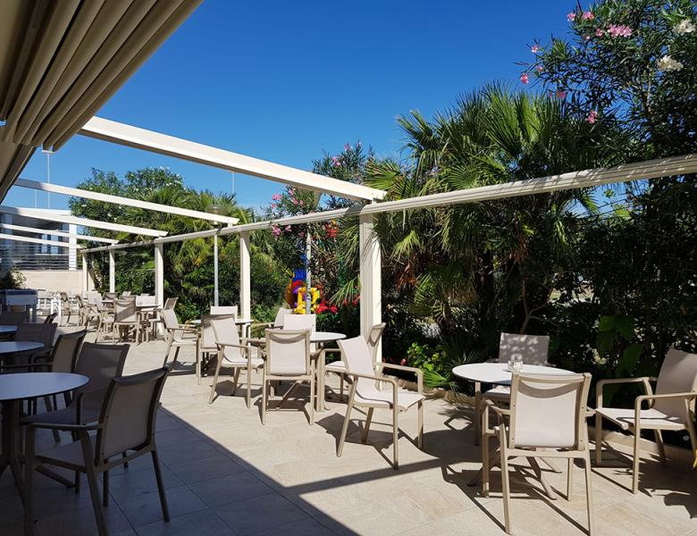 Terrazza soleggiata con sedie, tavoli e rigogliosa vegetazione tropicale.